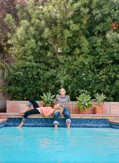 two people sitting on the edge of a swimming pool in front of some trees and bushes