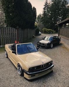 two cars are parked next to each other in the gravel near a fence and trees