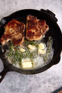 two pieces of meat are cooking in a skillet on the stove top with some herbs and butter