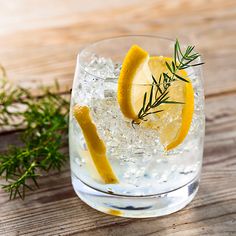 a glass filled with ice and lemon wedges on top of a wooden table next to an rosemary sprig