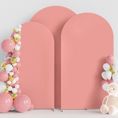 a teddy bear sitting next to some balloons on a pink wall and an arch shaped like a heart