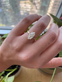 a woman's hand with a diamond ring on top of her finger, next to a potted plant