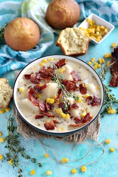 a bowl of soup with corn and bacon in it next to bread on a blue surface