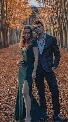 two people standing next to each other in front of trees with leaves on the ground