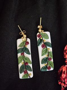 pair of earrings with green leaves and red berries on white ceramic rectangles, hanging from gold - plated ear wires