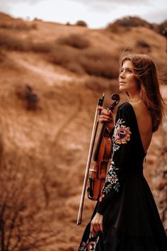 a woman in a black dress holding a violin and looking off into the distance while standing on a hill