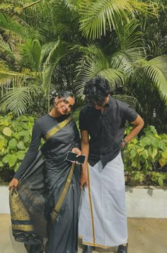 two women standing next to each other in front of some plants and trees with one woman wearing a black sari