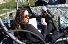 two people sitting in the snow with one holding a coffee cup and another taking a selfie