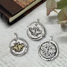 three silver and gold bee pendants sitting on top of a table next to a book