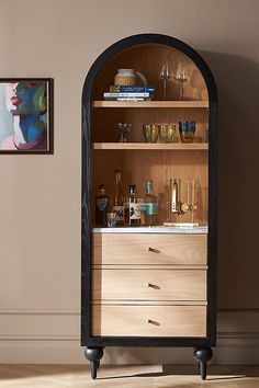 an arch shaped cabinet with wine glasses and liquor bottles on the top, in front of a painting