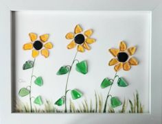 three sunflowers made out of sea glass in a white frame with green and yellow leaves