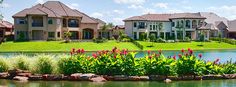 a pond surrounded by lush green grass next to two large houses with red flowers in the foreground