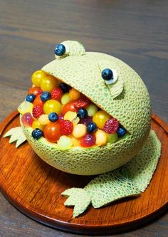 a frog made out of fruit sitting on top of a wooden plate with candy in it's mouth