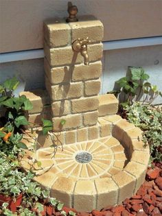 a small fountain in the middle of a flower bed next to a building with plants growing around it