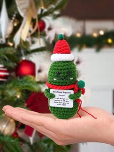 a hand holding a small crocheted christmas ornament in front of a tree