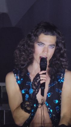 a woman with long curly hair holding a microphone in her right hand and wearing blue sequins