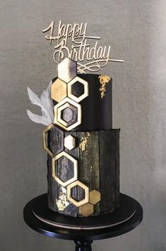 a black and gold birthday cake with white frosting on a pedestal in front of a gray wall