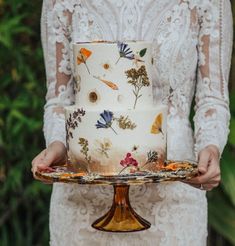 a woman in white dress holding a cake on a wooden platter with leaves and flowers