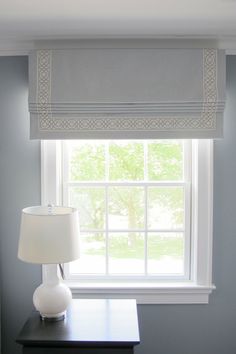 a white lamp sitting on top of a table next to a window with roman blinds