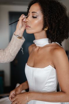 a woman is getting her make up done by another woman in a white dress and gold jewelry