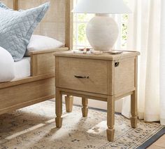 a nightstand with a lamp on top of it in front of a bed and window