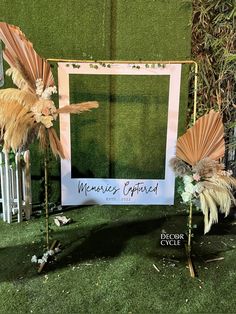 a photo frame with feathers and flowers on display in front of a green grass wall