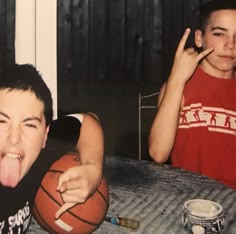 a young man sticking his tongue out next to a basketball on a table with another boy in the background