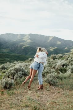 two women hugging each other in the middle of a field with mountains in the background