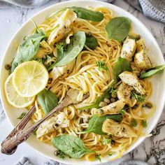 a white bowl filled with pasta and chicken on top of a marble table next to lemon wedges