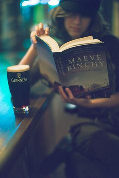 a woman sitting at a bar reading a book and drinking a pint of beer