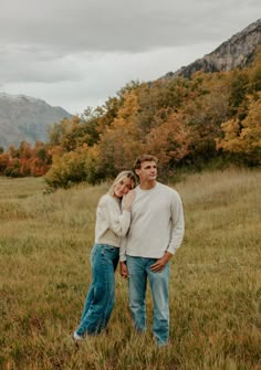 a man and woman standing in the grass with mountains in the background on an overcast day