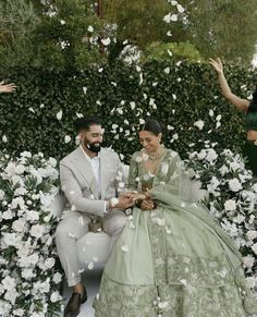 a man and woman dressed in green sitting on a bench surrounded by white flowers with petals flying around them