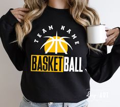 a woman is holding a coffee mug and wearing a black sweatshirt with the team name basketball on it