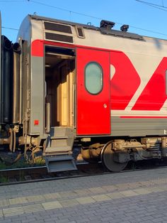 a red and white train car sitting on the tracks next to a brick sidewalk in front of a building