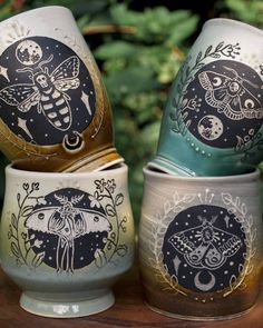 three painted egg cups sitting next to each other on top of a wooden table in front of trees