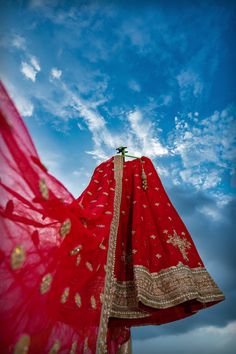 a woman wearing a red sari with gold accents on her head and arms, standing in front of a cloudy blue sky