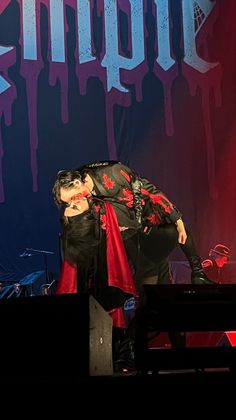 a male in a black shirt and red cape on stage with his hands over his head