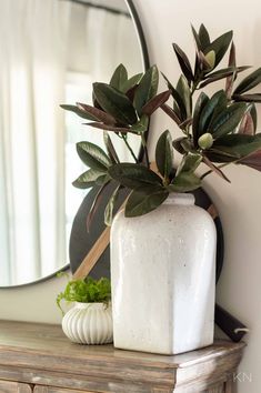 a white vase sitting on top of a wooden table next to a mirror and potted plant