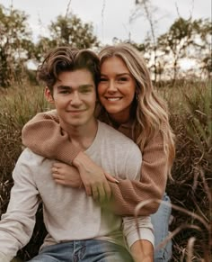 a young man and woman are sitting in tall grass smiling at the camera with their arms around each other