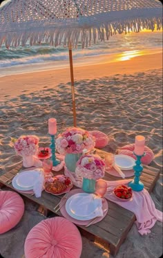 a table set up on the beach with plates and flowers in front of an umbrella