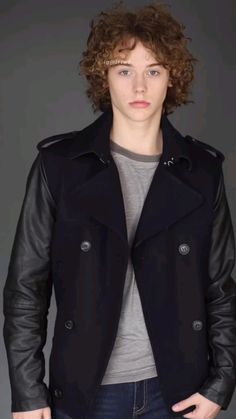 a young man with curly hair wearing a black jacket and grey t - shirt, standing in front of a gray background