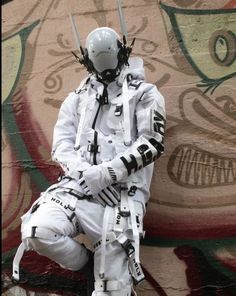 a man dressed in white sitting on the ground next to a wall with graffiti painted on it