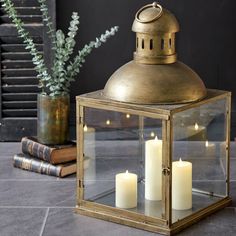 two lit candles in a brass lantern on the ground next to some books and a potted plant