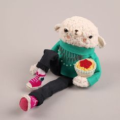 a white stuffed animal with a green sweater and pink shoes sitting on the ground holding a bowl of food