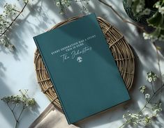 a green book sitting on top of a wicker basket next to white flowers and greenery