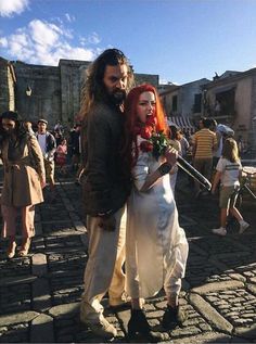 a man and woman dressed in medieval clothing standing on cobblestone street with people walking around