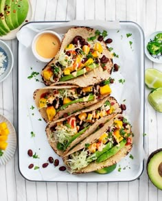 three tacos on a plate with salsa and avocado next to it, surrounded by other dishes