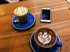 a cappuccino and cell phone sitting on a wooden table next to each other