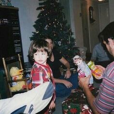 a group of people sitting around a christmas tree with presents on the floor and one person holding a doll