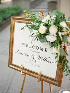 a welcome sign sitting on top of a wooden easel with flowers and greenery